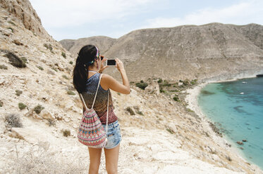 Rückansicht einer Frau mit Rucksack, die mit ihrem Smartphone das Meer fotografiert - CAVF14697