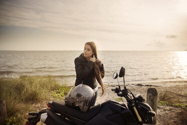 Junge Frau, die sich die Haare bindet, während sie mit einem Motorrad am Strand steht und auf das Meer blickt - CAVF14714