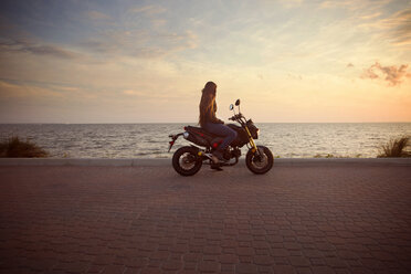 Seitenansicht der Frau fährt Motorrad auf der Straße gegen das Meer bei Sonnenuntergang - CAVF14722