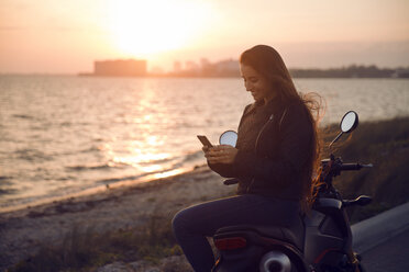 Glückliche Frau, die ein Smartphone benutzt, während sie bei Sonnenuntergang am Meer sitzt - CAVF14723