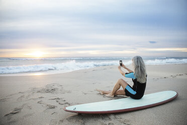 Seitenansicht eines Surfers, der mit seinem Smartphone fotografiert, während er auf dem Surfbrett am Strand von Delray sitzt - CAVF14730