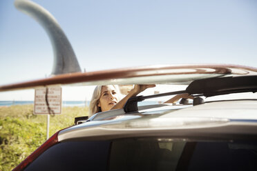 Mature woman tying surfboard on car roof at Delray beach against clear blue sky - CAVF14745