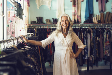 Porträt einer selbstbewussten Besitzerin, die lächelnd an einem Kleiderständer im Geschäft steht - CAVF14757