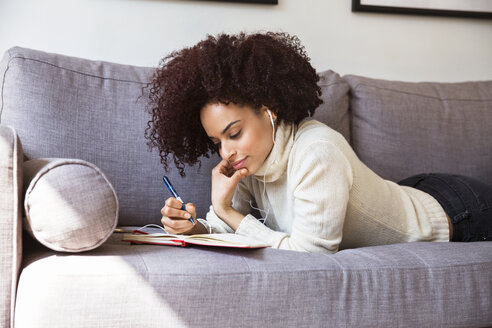 Frau schreibt in einem Buch, während sie auf dem Sofa Musik hört - CAVF14773