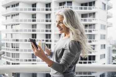 Happy mature woman using smart phone while holding coffee mug on balcony against building - CAVF14775
