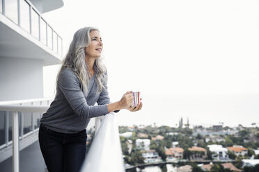 Thoughtful mature woman holding coffee mug while standing on balcony - CAVF14779