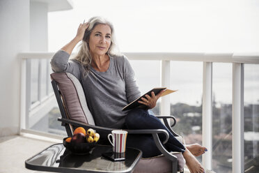 Porträt einer selbstbewussten Frau, die ein Buch hält und auf einem Stuhl auf dem Balkon sitzt - CAVF14784