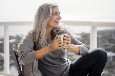 Nachdenkliche reife Frau, die eine Kaffeetasse hält, während sie auf dem Balkon sitzt - CAVF14787