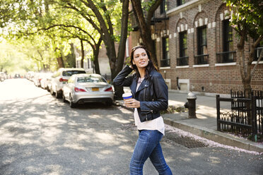 Young woman holding disposable cup while crossing city street - CAVF14811