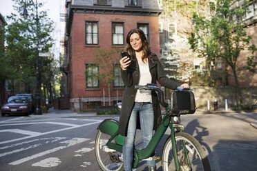 Junge Frau benutzt ihr Smartphone, während sie mit dem Fahrrad auf der Straße steht - CAVF14830