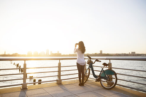 Rückansicht einer Frau, die die Stadt fotografiert, während sie mit dem Fahrrad auf der Promenade bei Sonnenuntergang steht - CAVF14832