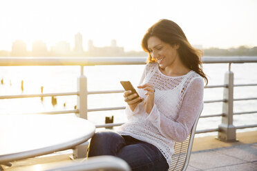 Happy woman using smart phone at cafe on promenade during sunset - CAVF14833