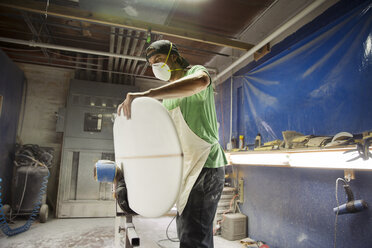 Worker holding surfboard while standing in workshop - CAVF14845