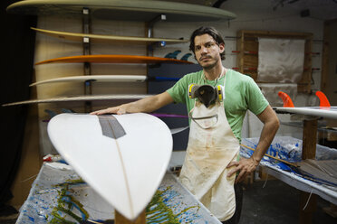 Porträt eines ernsthaften männlichen Arbeiters, der in einer Werkstatt neben einem Surfbrett steht - CAVF14853