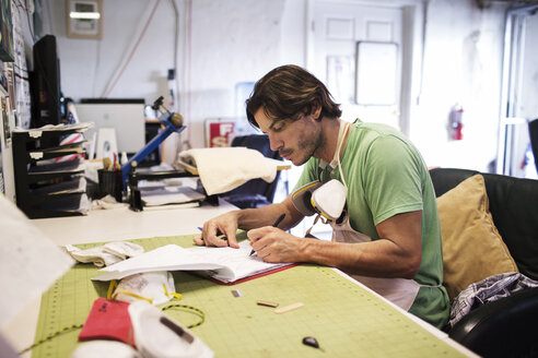 Mann schreibt auf Papier, während er am Tisch in einer Werkstatt sitzt - CAVF14854