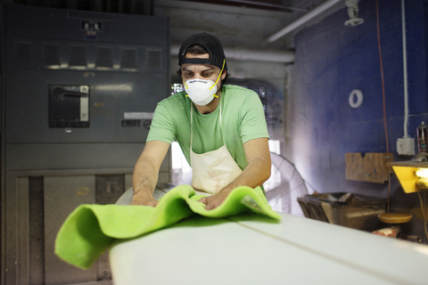 Männlicher Arbeiter, der in einer Werkstatt ein Surfbrett mit Stoff abwischt, lizenzfreies Stockfoto