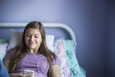 Lächelndes Mädchen, das ein Smartphone benutzt, während es sich auf dem Bett entspannt - CAVF14953