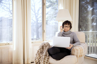 Ältere Frau benutzt Smartphone und Laptop, während sie zu Hause auf einem Sessel sitzt - CAVF14969