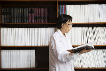 Senior female scientist reading book by shelves at laboratory - CAVF15006