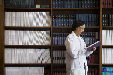 Side view of senior female scientist reading book by shelves at laboratory - CAVF15008