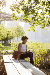 Woman using smart phone while sitting on bench in park - CAVF15042