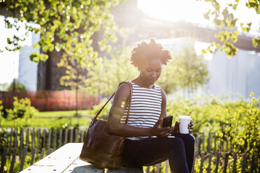 Woman using smart phone while sitting on bench in park during summer - CAVF15043