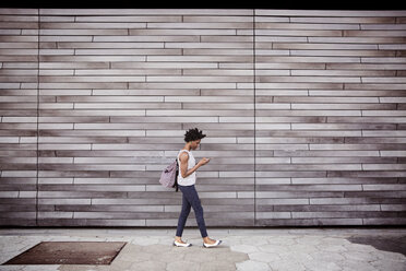 Side view of woman using smart phone while walking against wall - CAVF15049