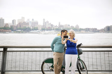 Cheerful mature couple taking selfie on promenade by river in city - CAVF15061