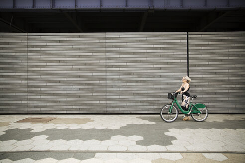 Seitenansicht einer reifen Frau, die mit dem Fahrrad auf dem Fußweg an der Mauer spazieren geht - CAVF15064