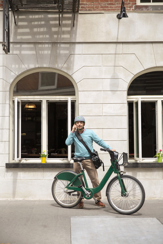 Älterer Mann, der ein Smartphone benutzt, während er mit dem Fahrrad vor einem Gebäude steht, lizenzfreies Stockfoto
