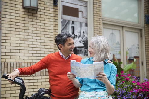 Happy mature couple reading road map while standing with bicycle outside building - CAVF15072