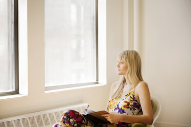 Nachdenkliche Geschäftsfrau, die ein Buch und einen Stift hält, während sie auf einem Sessel in einem kreativen Büro sitzt - CAVF15080