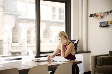 Businesswoman reading file at desk by window in creative office - CAVF15092