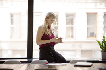 Geschäftsfrau, die ein Smartphone benutzt, während sie auf der Fensterbank im Sitzungssaal sitzt - CAVF15095