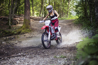 Female biker riding dirt bike on puddle in forest - CAVF15236