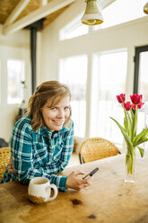 Porträt einer Frau, die ein Smartphone benutzt, während sie zu Hause am Tisch sitzt - CAVF15312