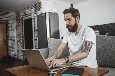 Bärtiger Mann sitzt auf der Couch zu Hause und benutzt einen Laptop - VPIF00396