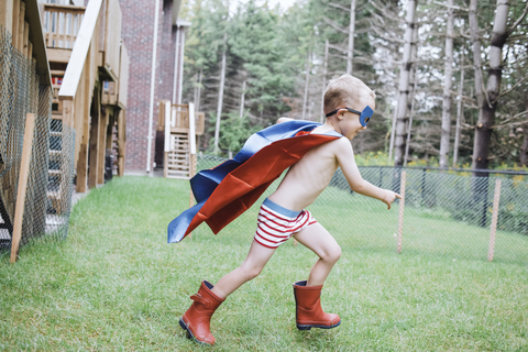 Seitenansicht eines hemdlosen Jungen mit Umhang und Augenklappe beim Spielen im Garten, lizenzfreies Stockfoto