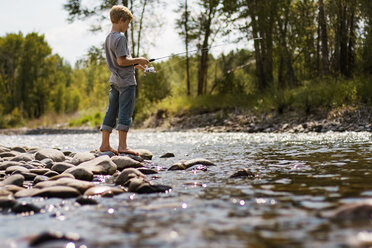 Side view of boy fishing while standing on rocks in river at forest - CAVF15378