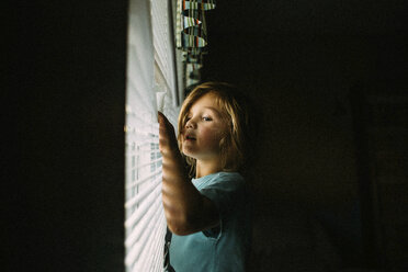 Mädchen schaut durch die Jalousien, während sie in der Dunkelkammer zu Hause steht - CAVF15382