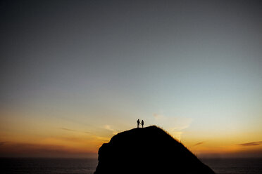 Distant view of couple standing on silhouette cliff against sky by sea during sunset - CAVF15424