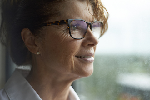 Frau mit Sonnenbrille schaut weg, während sie zu Hause am Fenster steht, lizenzfreies Stockfoto