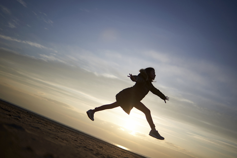 Glückliches Mädchen springt am Strand gegen den Himmel bei Sonnenuntergang, lizenzfreies Stockfoto