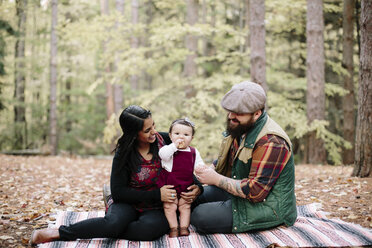 Glückliche Eltern sehen ihre Tochter beim Essen an, während sie sich auf einer Decke im Wald entspannen - CAVF15553