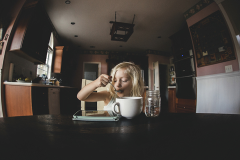 Mädchen isst Essen und schaut auf einen Tablet-Computer auf dem Tisch, lizenzfreies Stockfoto