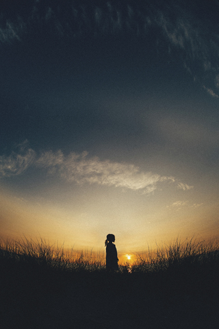 Silhouette Mädchen stehend auf grasbewachsenen Feld gegen den Himmel bei Sonnenuntergang, lizenzfreies Stockfoto