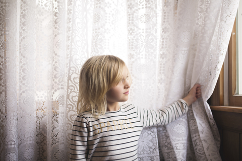 Nachdenkliches Mädchen, das vor einem Vorhang steht und durch ein Fenster schaut, lizenzfreies Stockfoto