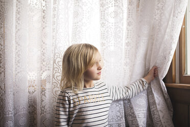 Thoughtful girl standing against curtain looking through window - CAVF15639