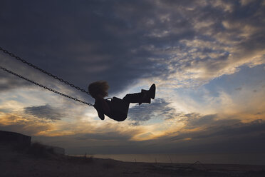 Side view of girl playing on swing at beach against cloudy sky during sunset - CAVF15645