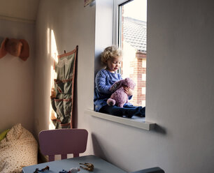 Mädchen spielt mit Stofftier, während sie zu Hause auf der Fensterbank sitzt - CAVF15656